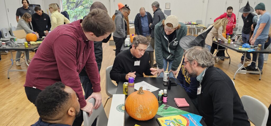 A group of people is gathered around a table, collaborating on what seems to be the design or planning phase of their pumpkin racer project for a team building event. There is a pumpkin on the table, along with various crafting supplies such as glue, markers, and construction paper. The participants appear engaged in a discussion, possibly strategizing or finalizing the design for their pumpkin racer.