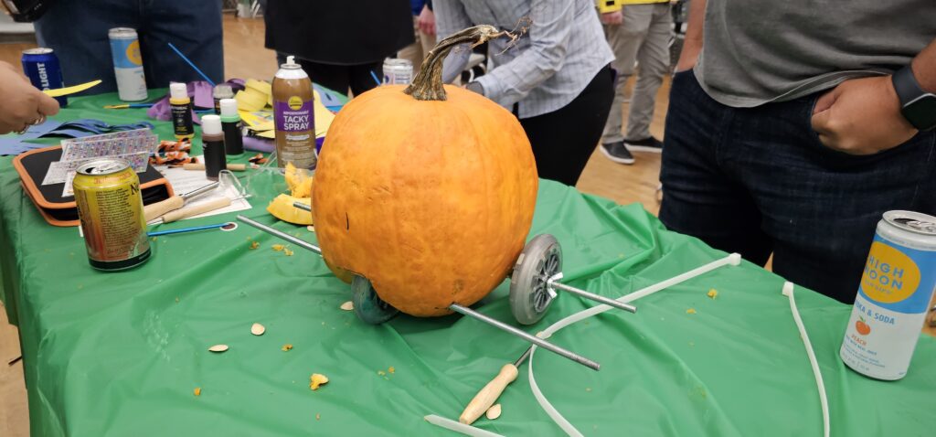 This image shows a pumpkin racer in the process of being assembled during a team building event. The pumpkin is fitted with metal rods serving as axles, and wheels are attached on either side. The scene captures the creativity of participants as they construct their racer, likely in preparation for a fun pumpkin race. The surrounding materials, drinks, and craft supplies add to the lively and collaborative atmosphere.