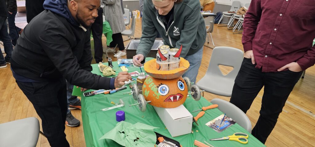 A group of individuals is working on a pumpkin racer, a fun Halloween-themed activity. The pumpkin racer has large, googly eyes and a wide mouth with pointed teeth painted on it. It is placed on wheels, ready for racing. The table is covered in tools and decorations such as scissors and crafting materials, while the team building participants focus on adding final touches to the pumpkin racer.