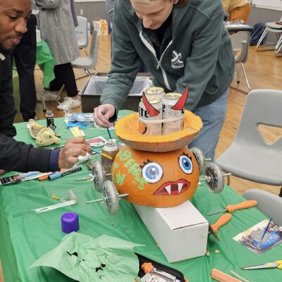 A group of individuals is working on a pumpkin racer, a fun Halloween-themed activity. The pumpkin racer has large, googly eyes and a wide mouth with pointed teeth painted on it. It is placed on wheels, ready for racing. The table is covered in tools and decorations such as scissors and crafting materials, while the team building participants focus on adding final touches to the pumpkin racer.