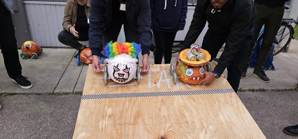 Two individuals are preparing pumpkin-themed cars for a race on a wooden surface. The pumpkin on the left is decorated with a colorful clown wig and a painted face, while the pumpkin on the right features a more monster-like design with sharp teeth and attached eyes. Both pumpkins have wheels attached, ready to race. A checkerboard line is visible on the board, marking the starting point of this Great Pumpkin Team Building Race.