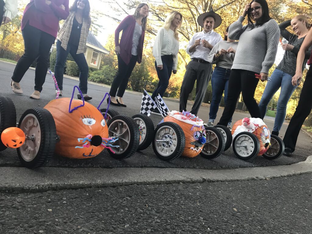 This image captures a fun moment from a pumpkin team building race event. Three creatively decorated pumpkins are lined up, each mounted on wheels to form pumpkin racers. The racers have unique designs: one appears to have been styled like a cat, with pipe cleaner whiskers and ears. Others are adorned with checkered flags and various festive decorations. In the background, participants are smiling and enjoying the activity, clearly excited about the upcoming race.