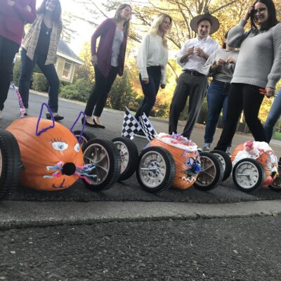 This image captures a fun moment from a pumpkin team building race event. Three creatively decorated pumpkins are lined up, each mounted on wheels to form pumpkin racers. The racers have unique designs: one appears to have been styled like a cat, with pipe cleaner whiskers and ears. Others are adorned with checkered flags and various festive decorations. In the background, participants are smiling and enjoying the activity, clearly excited about the upcoming race.