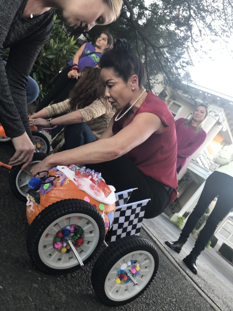 A participant is in the middle of a pumpkin race team building event. The pumpkin racer is decorated with colorful pom-poms, pipe cleaners, and a miniature checkered flag, symbolizing the competitive yet festive atmosphere of the race. The person is focused on adjusting the wheels of the creatively designed pumpkin racer, which is fitted with large wheels for mobility.