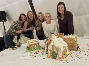 Five smiling participants gather around a table displaying their decorated gingerbread houses during a team building event. The gingerbread houses are intricately adorned with icing, candy, and festive decorations, including a snowman figure on one of the houses.