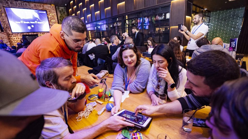 Team members huddle around their iPad, strategizing and collaborating to solve various puzzles, surrounded by game materials like hacky sacks and play dough. The fast-paced, interactive environment fuels teamwork and creative thinking.