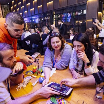 Team members huddle around their iPad, strategizing and collaborating to solve various puzzles, surrounded by game materials like hacky sacks and play dough. The fast-paced, interactive environment fuels teamwork and creative thinking.