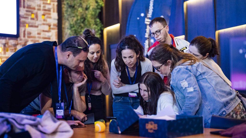 A group of participants collaborates around a tablet, solving challenges together during an interactive team building event.