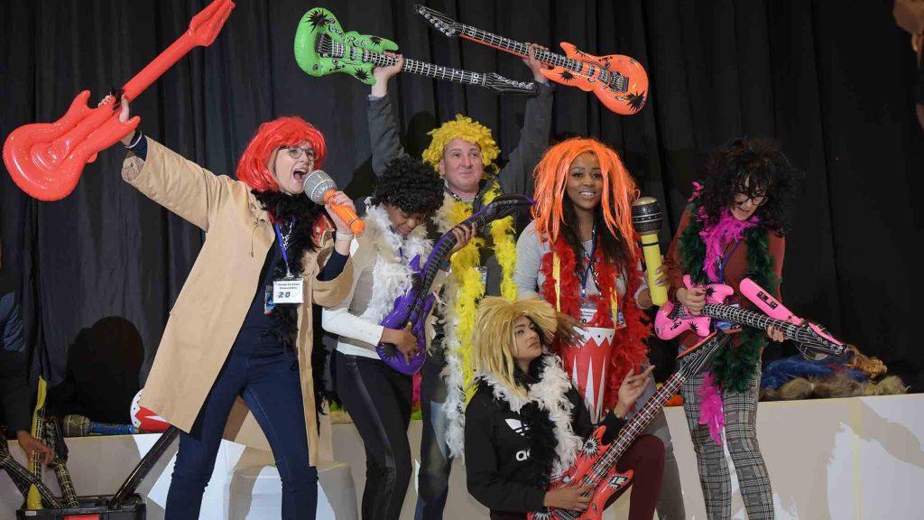 A group of participants dressed in colorful costumes and holding inflatable guitars celebrates with enthusiasm during a team building event, showcasing their creativity and energy. Their spirited performance highlights the fun atmosphere of the activity.