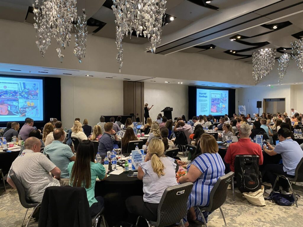 Participants at a large Hopefull Handbags Global team building event, listening to a speaker as they prepare to pack handbags with essentials for domestic abuse survivors.