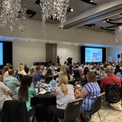 Participants at a large Hopefull Handbags Global team building event, listening to a speaker as they prepare to pack handbags with essentials for domestic abuse survivors.