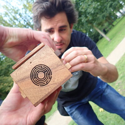 Participant carefully examines a small wooden puzzle box with a maze design during the 'Escape The Maze' team building event, focusing on solving the challenge.