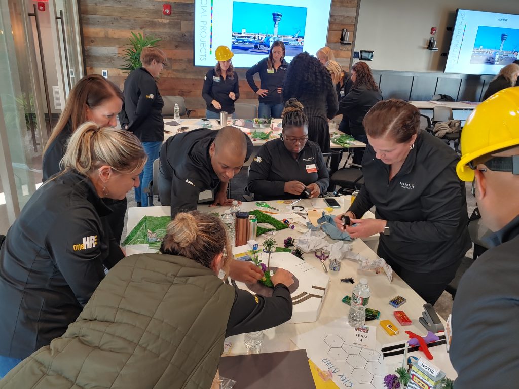 Team members collaborating on a City Build team building project, working together to construct miniature city zones using materials, models, and creative elements.