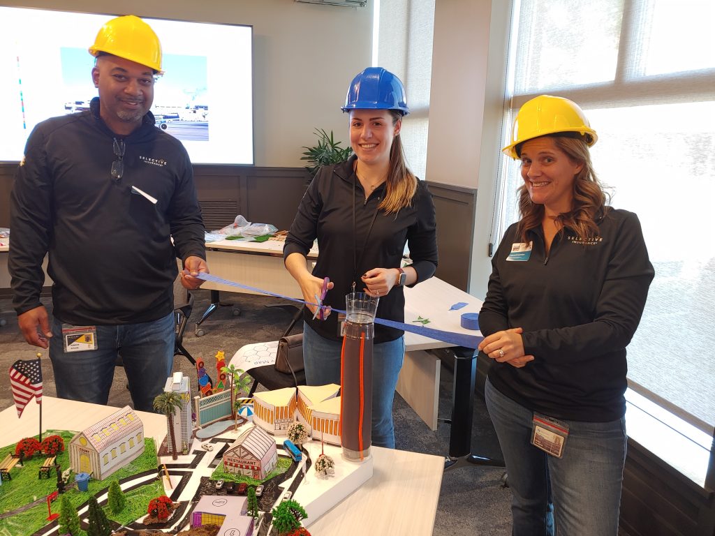 Three team members in hard hats proudly cutting a ribbon to unveil their collaborative City Build project, featuring a detailed miniature urban landscape.