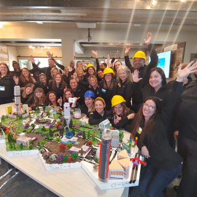 A large group of participants celebrates their success at a City Build team building event, posing around a detailed miniature city model while wearing hard hats and smiling enthusiastically.