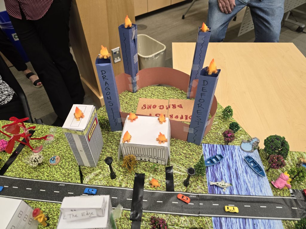 Creative city model with labeled towers, roads, waterways, and miniature vehicles, showcasing teamwork from the City Build team building event.