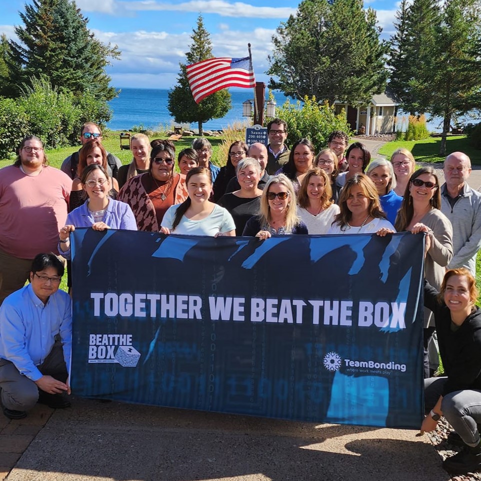 Group of colleagues proudly pose with a 'Together We Beat the Box' banner, celebrating their teamwork and success in the Beat the Box team building challenge.