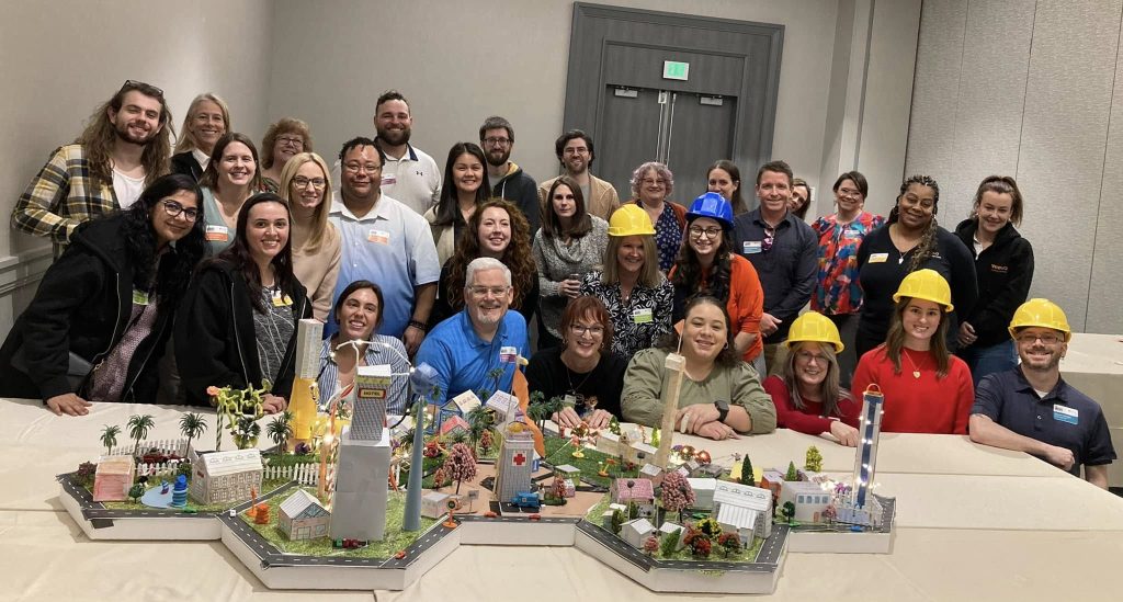 Group photo of participants wearing hard hats, proudly posing behind their completed miniature city during a City Build team building event.