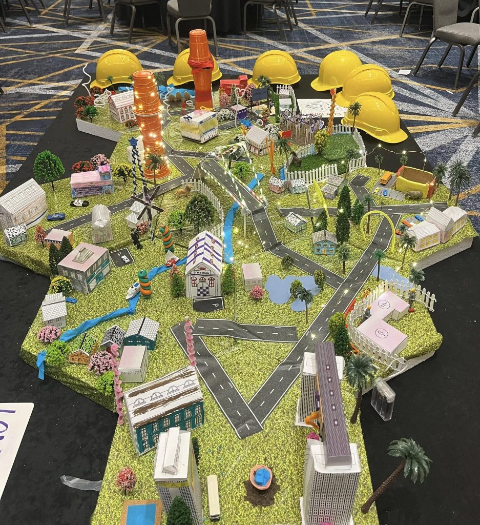 A completed City Build team building project featuring a vibrant miniature city with buildings, roads, trees, and glowing lights, surrounded by yellow hard hats on display.