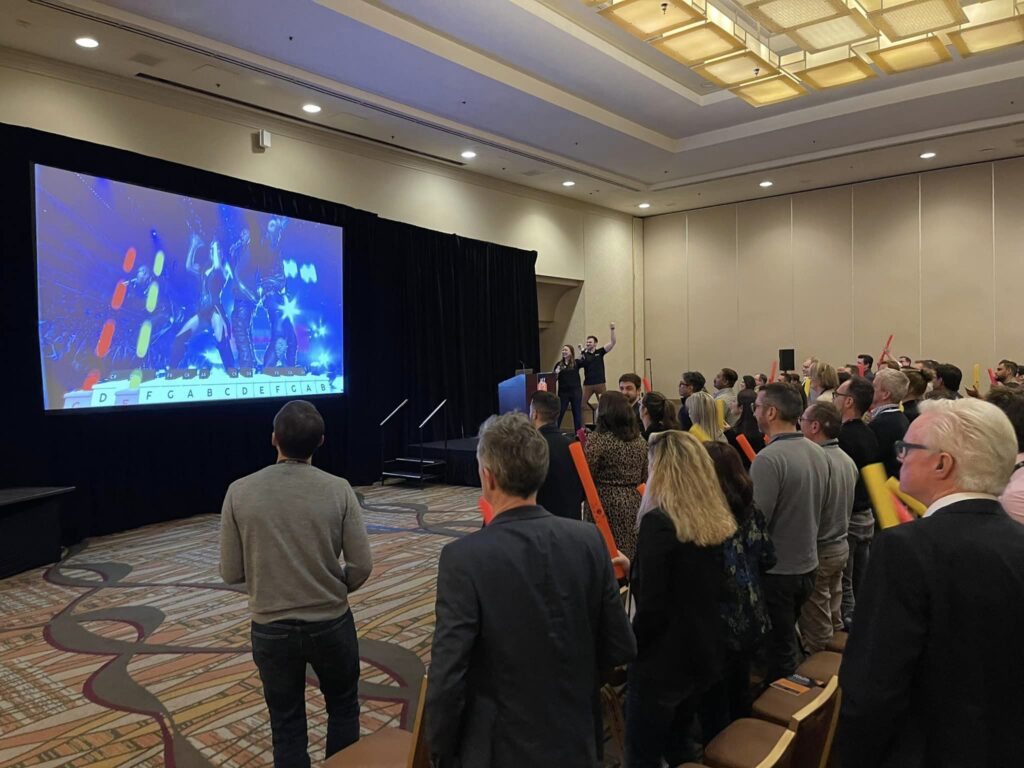 Participants in a Boom Time team building event are actively following a colorful rhythm-based music display projected on a large screen. Holding Boomwhackers, they collaborate to play the correct notes, guided by the display, in a fun and engaging team activity.