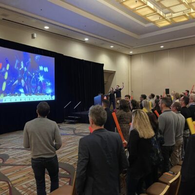 Participants in a Boom Time team building event are actively following a colorful rhythm-based music display projected on a large screen. Holding Boomwhackers, they collaborate to play the correct notes, guided by the display, in a fun and engaging team activity.