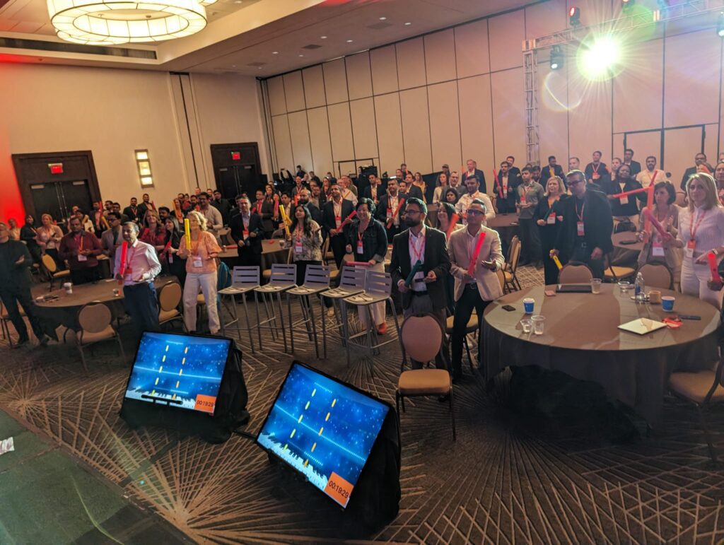 Conference participants engage in a high-energy Boom Time team building session, using colorful Boomwhackers to follow musical patterns displayed on large screens. The event promotes collaboration, rhythm, and excitement in a lively setting.