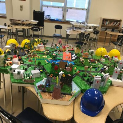 Colorful miniature city model displayed on tables, surrounded by yellow and blue hard hats, featuring buildings, roads, and greenery for a City Build team building activity.