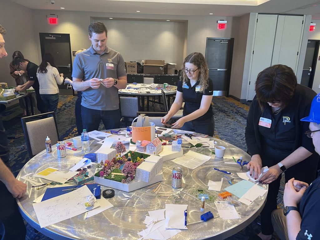 Team members at a City Build team building event collaborate to design and assemble a creative miniature city model using various materials on a round table.