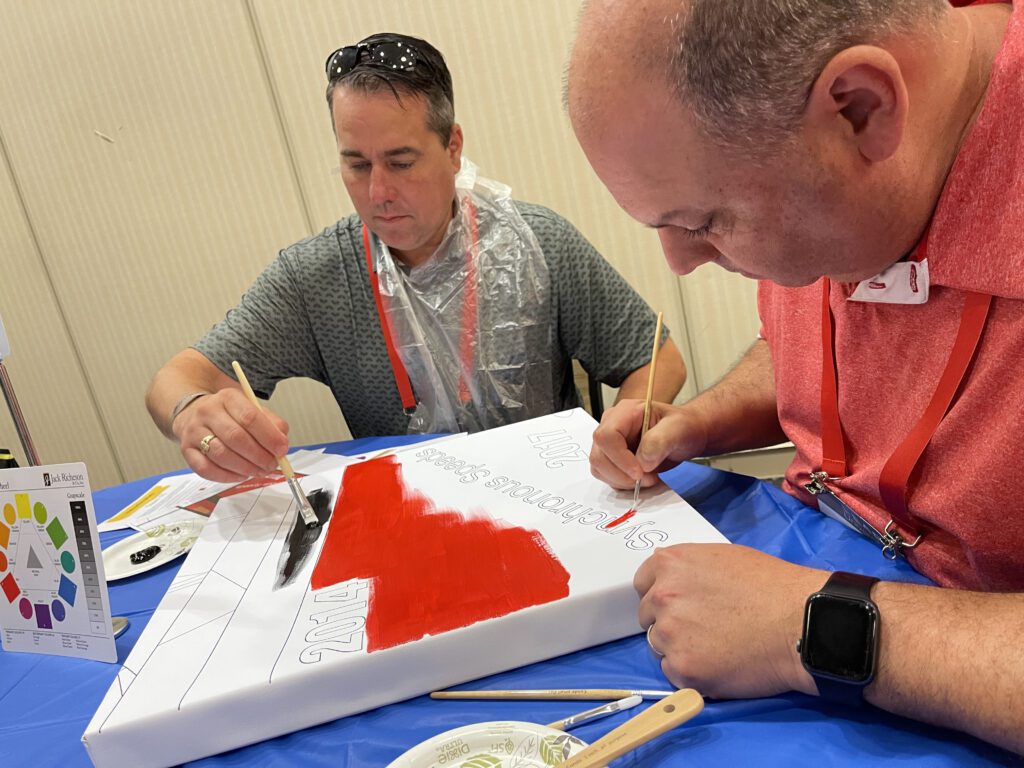 Two men carefully paint a section of a mural during The Big Picture team building event. They are focused on adding red and black colors to their canvas, with a color wheel and brushes nearby.