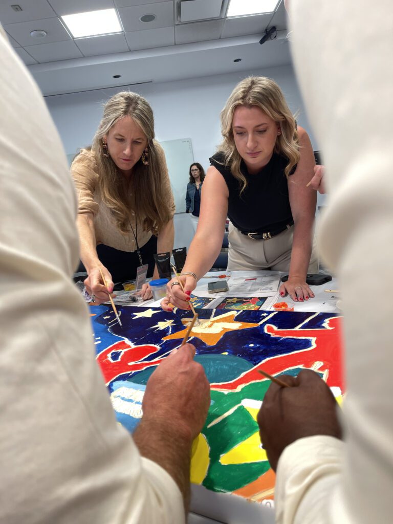 Two team members concentrate as they add final touches to their section of a vibrant mural during The Big Picture team building event, fostering collaboration and creativity.