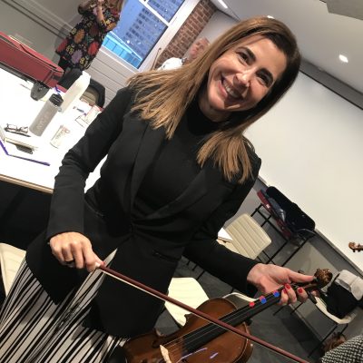 Smiling participant holds a violin and bow during the Crescendo team building event, standing in a meeting room with tables and chairs.