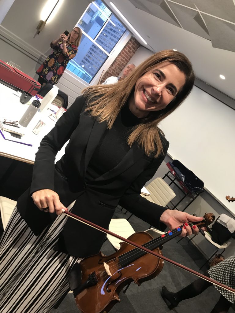 Smiling participant holds a violin and bow during the Crescendo team building event, standing in a meeting room with tables and chairs.