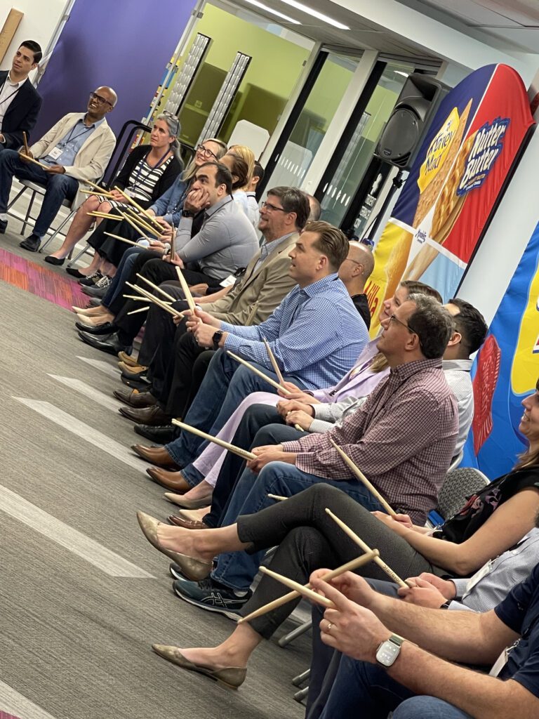 A group of participants seated in a row, holding drumsticks and preparing for a rhythm-based activity during a BeatsWork team building event. Everyone is focused and ready to engage in the upcoming musical challenge.