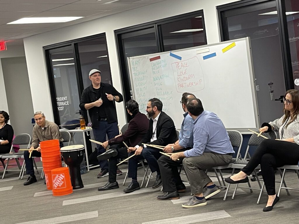 A facilitator leads a drumming session during a BeatsWork team building event, with participants seated in a semi-circle, holding drumsticks. A whiteboard with notes is visible in the background, setting the rhythm for the engaging musical activity.