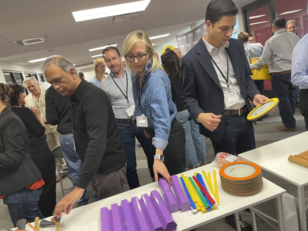 Participants select percussion instruments from a table during a BeatsWork team building event, preparing to engage in rhythm-based activities. The group is focused on choosing colorful tubes and drums for their upcoming performance.