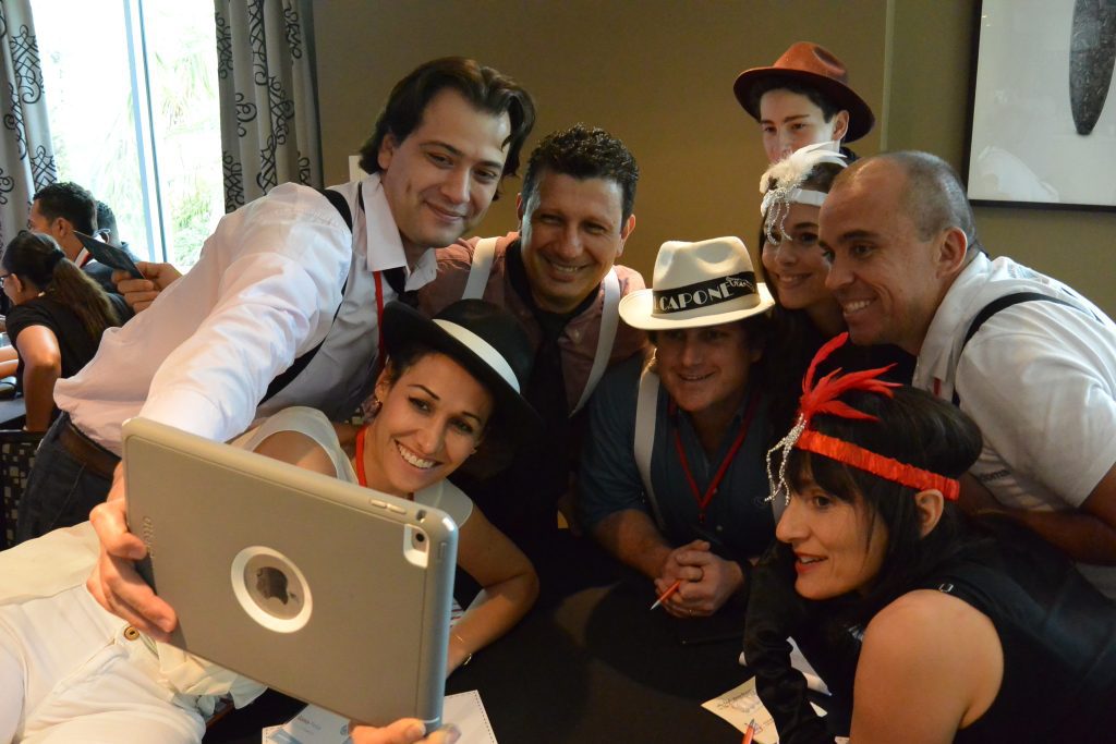 A team dressed in 1920s gangster-themed outfits gathers for a selfie during the high-energy Escape the Mob team building event, solving clues and creating their alibi.