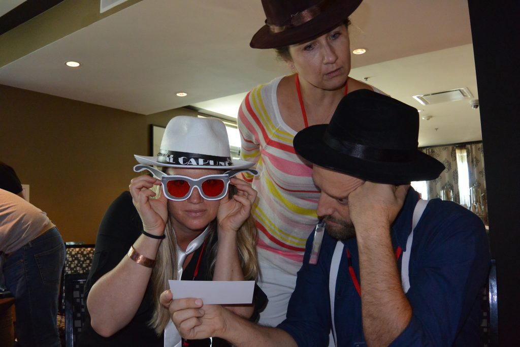 A team participating in the Escape the Mob event focuses on solving clues, while wearing fun mobster-themed hats and sunglasses to fit the robbery mystery challenge.