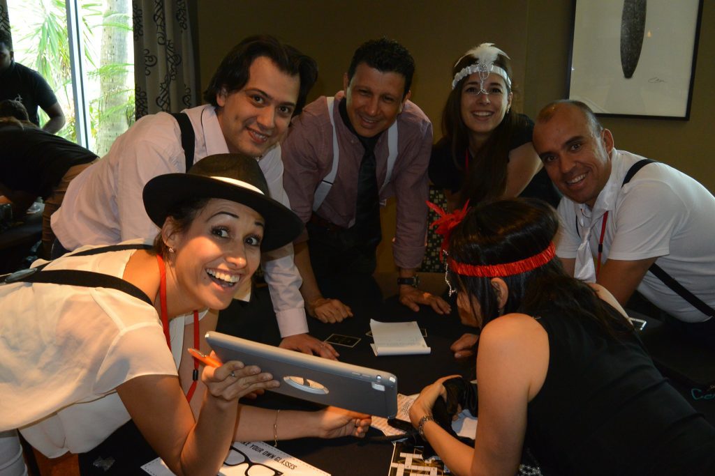 A group of participants in the Escape the Mob team building event works together, using a tablet to solve clues and track down the stolen diamonds while embracing their mobster-themed costumes.