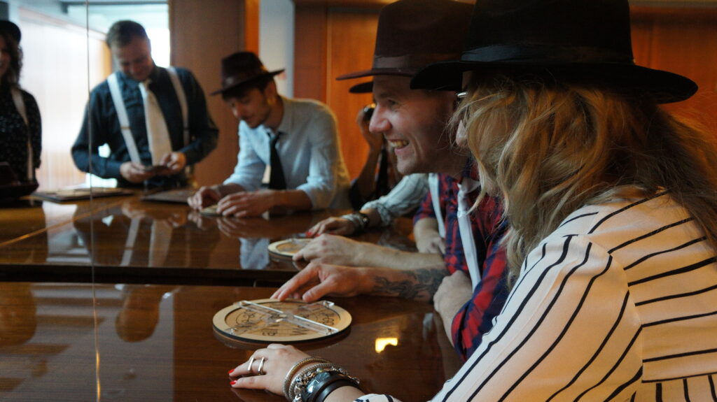 Group of participants dressed in vintage-style clothing, including hats, enthusiastically engaging in the 'Escape the Mob' team building activity. They are gathered around a table, analyzing a round puzzle piece, showing excitement and teamwork to solve the mystery.