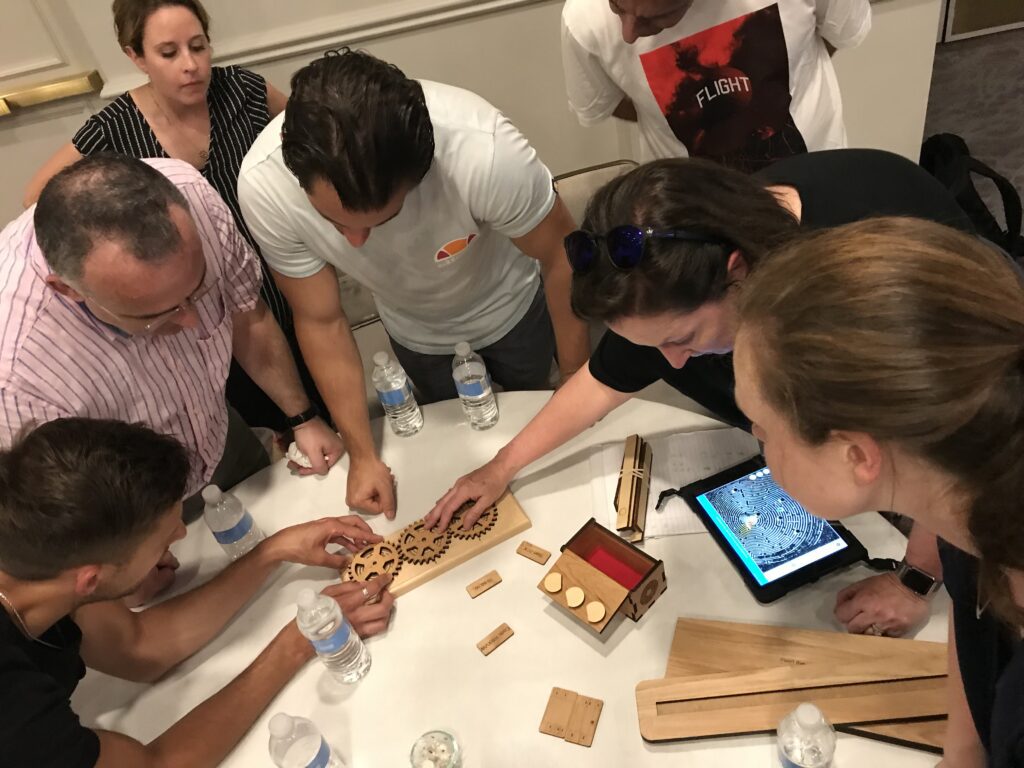 Team collaborates on a mechanical puzzle at a table, working together to solve a challenge in the 'Escape The Maze' team building activity.