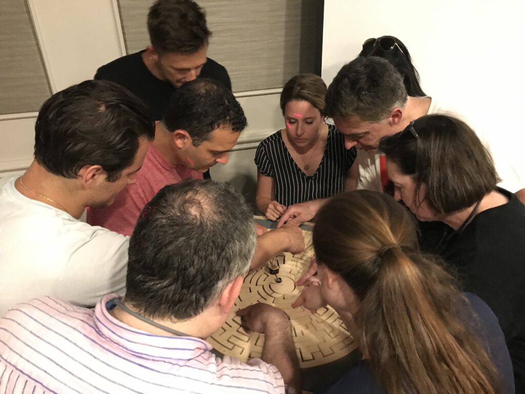 Team collaborates intently around a wooden maze puzzle during the 'Escape The Maze' team building challenge, focusing on strategy and teamwork.