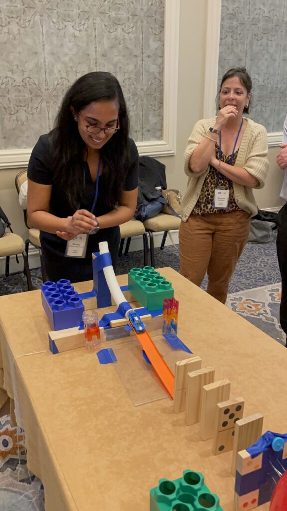 Participants eagerly watch a chain reaction contraption in action during a team building activity. A woman smiles as she sets off the colorful domino and building block setup on the table.