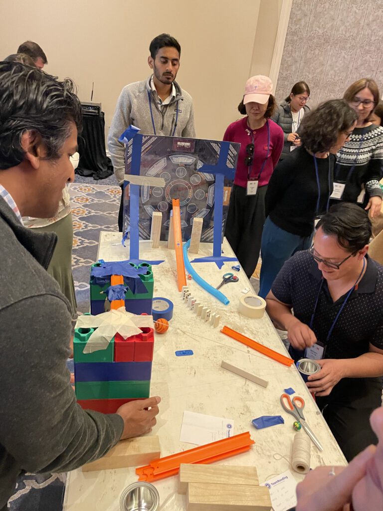 Participants collaborate to build a Rube Goldberg-inspired machine for a chain reaction team building activity, using colorful blocks, tracks, and other materials.