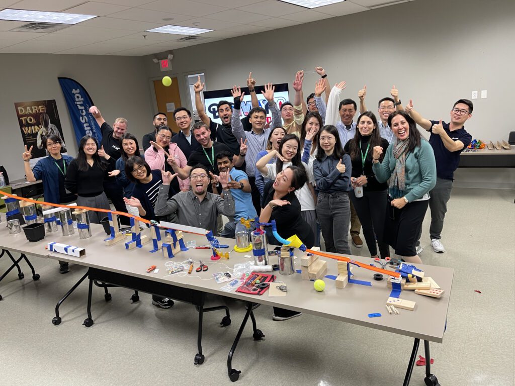 A group of excited participants celebrate their collaborative success after completing a Rube Goldberg-inspired contraption during the Domino Effect team building event.