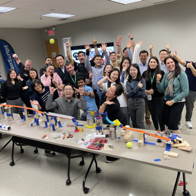 A group of excited participants celebrate their collaborative success after completing a Rube Goldberg-inspired contraption during the Domino Effect team building event.