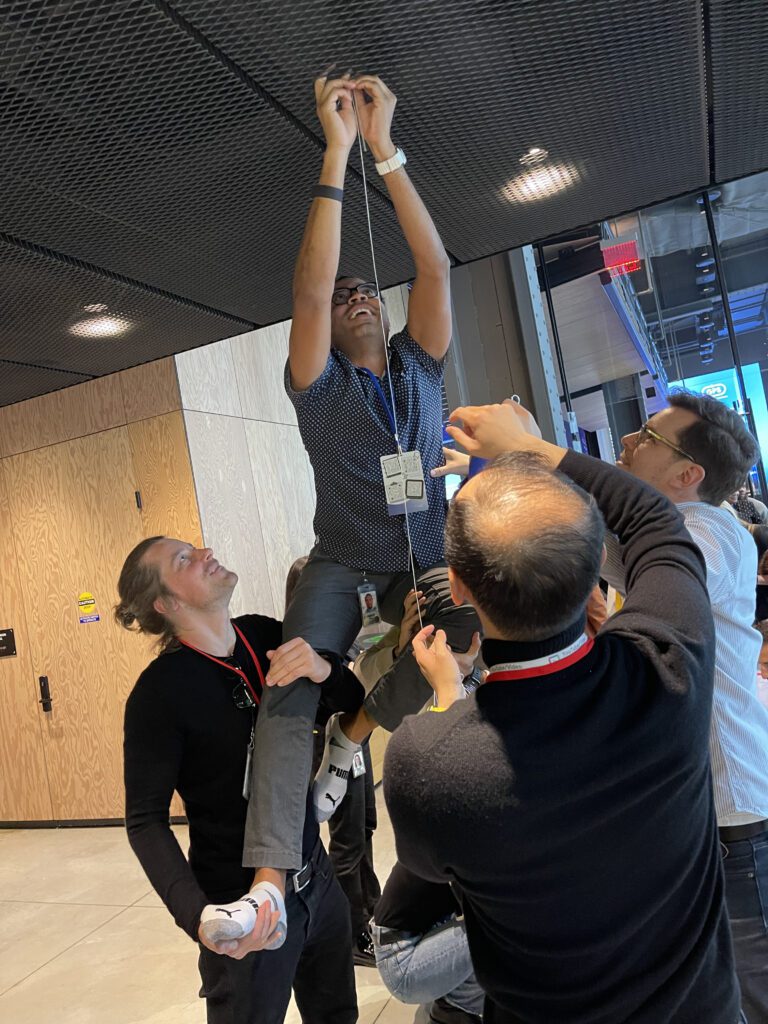 Team members work together to lift a colleague, helping him reach up to complete a part of their chain reaction machine in a collaborative team building exercise.