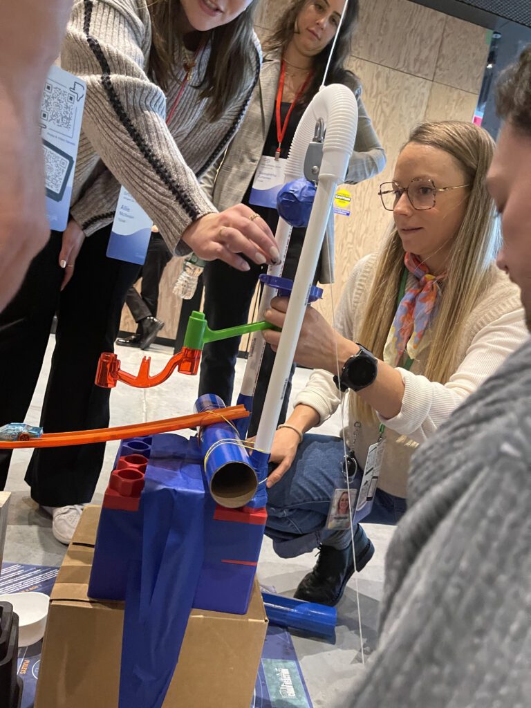 Participants carefully assemble pieces for a Rube Goldberg-inspired machine during a Chain Reaction team building activity, focusing on intricate connections and design.