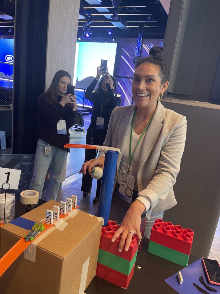 Participant excitedly tests her team's chain reaction machine at a team building event, with others capturing the moment in the background.