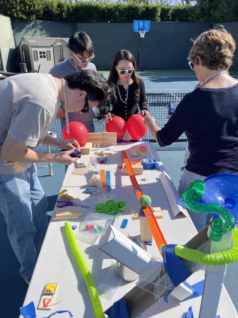 Team collaborating outdoors to assemble a creative Rube Goldberg-style machine during the Domino Effect team building activity.