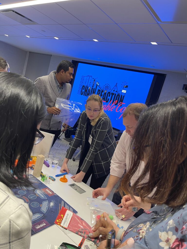 Team members collaborate intensely on their chain reaction machine during a team building event, with a 'Chain Reaction' sign displayed in the background.